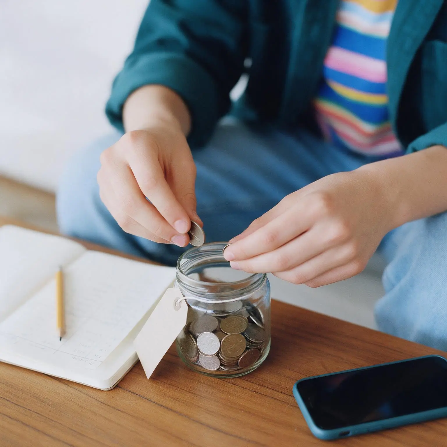 Unas manos echando monedas en un frasco de ahorro junto a un cuaderno.